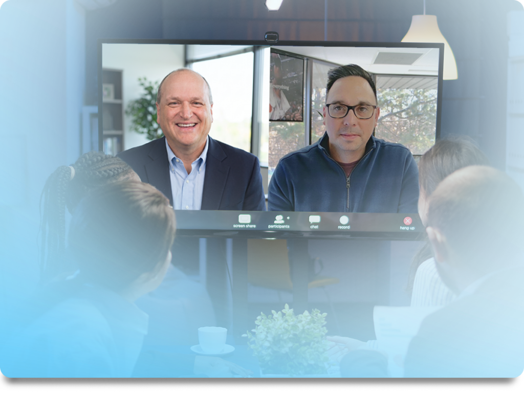 Video conference screen showing two men on a call; attendees sit around a table in the foreground with coffee and a plant visible on the table.